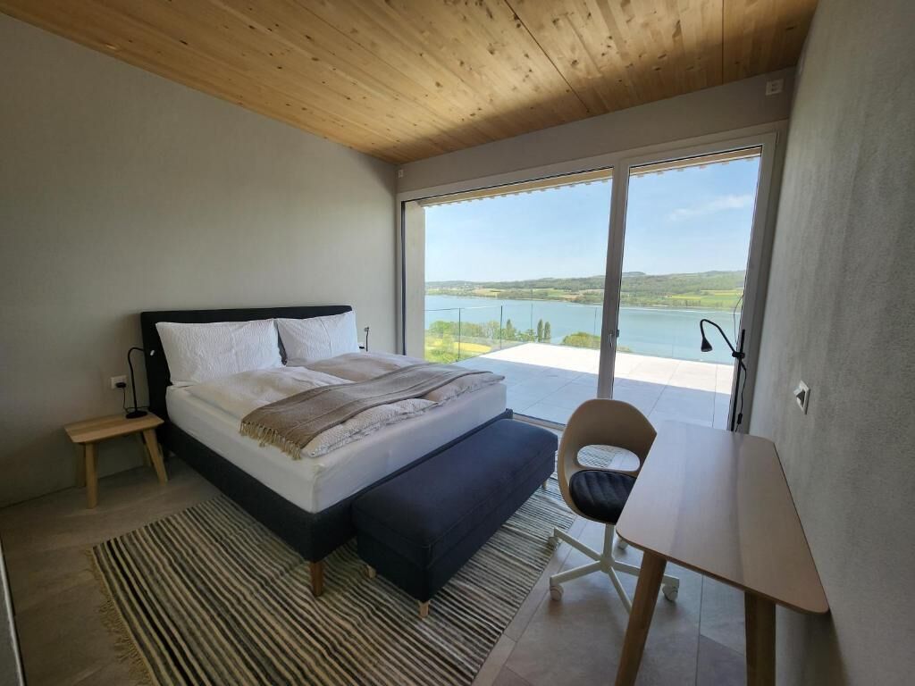 A bedroom with a bed, a chair, a small table, a view of a lake, and a wooden ceiling.