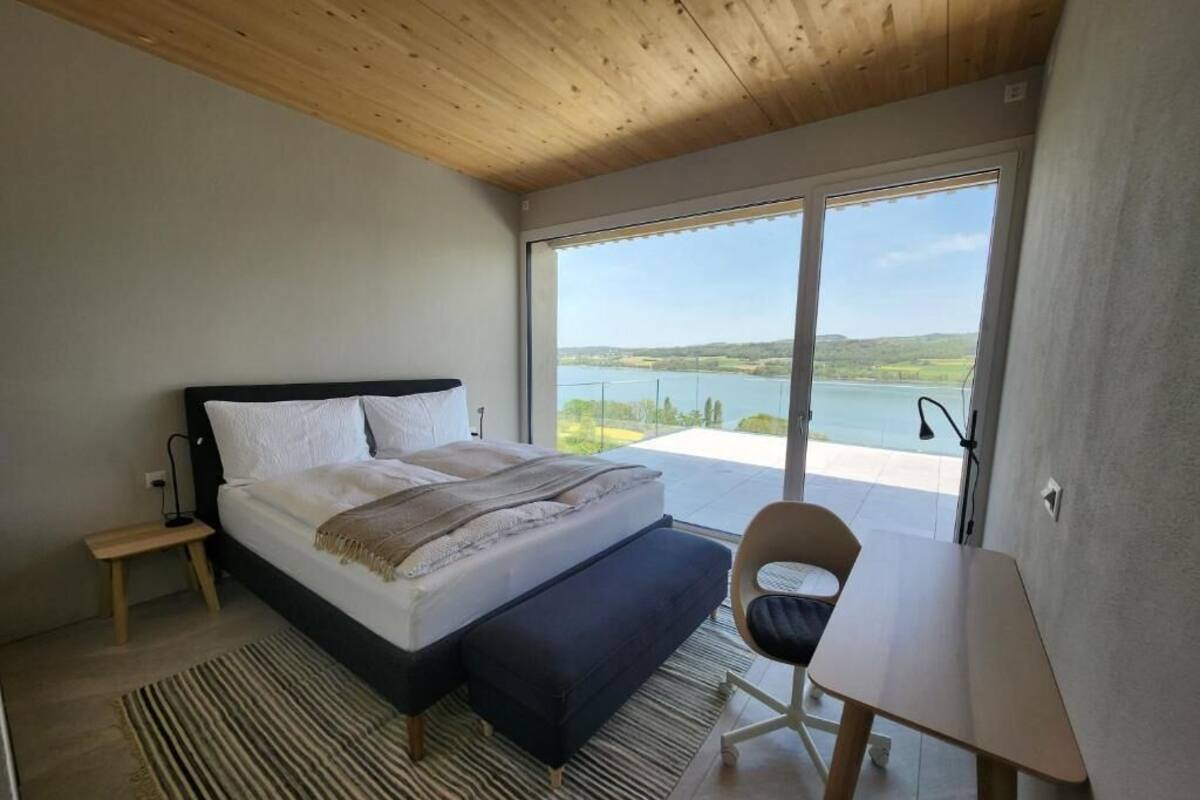 A bedroom with a bed, a chair, a small table, a view of a lake, and a wooden ceiling.