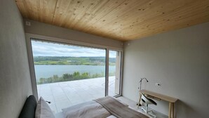 A bedroom with a large window, a bed, a desk, and a view of a lake.
