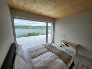 A bedroom with a large window, a bed, a desk, and a view of a lake. - Lakeview Guest Rooms Rooftop Terrace (Ebikon)