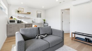 A modern living room with a grey sofa, a coffee machine, and a small kitchen area with a microwave and sink.