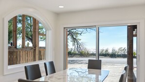 A dining area with a large table and chairs, a view of the beach through a glass door, and a wooden deck outside.