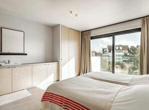 Een moderne slaapkamer met een groot bed, een minimalistische badkamer en een balkon met uitzicht op gebouwen.