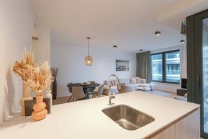 A modern kitchen with a central island, stainless steel sink, and a dining area with a table and chairs. - Modern apartment in the Iconic Ensor Tower (Oostende)