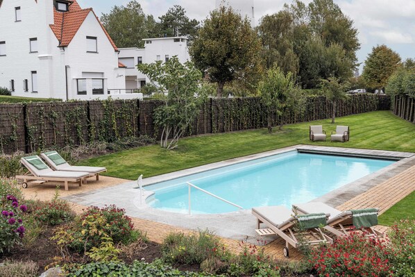 A rectangular swimming pool with a wooden deck, surrounded by a well-maintained lawn and a variety of flowering plants.