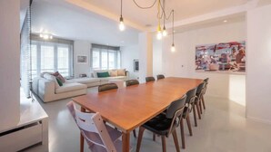 Ein modernes Wohnzimmer mit einem großen Esstisch aus Holz, einer Couch und einer Küchenzeile mit großem Fenster.