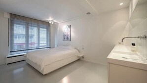 Ein modernes Schlafzimmer mit Bett, einem Fenster mit Jalousien, einem Wandbild und einem Badezimmer mit Waschbecken und Spiegel.