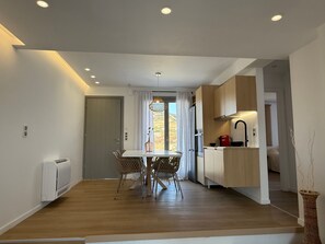 A modern kitchen with wooden cabinets, a dining area with a table and chairs, and a view through a window with curtains. - 2-bedroom villa Odysseus (Antiparos)