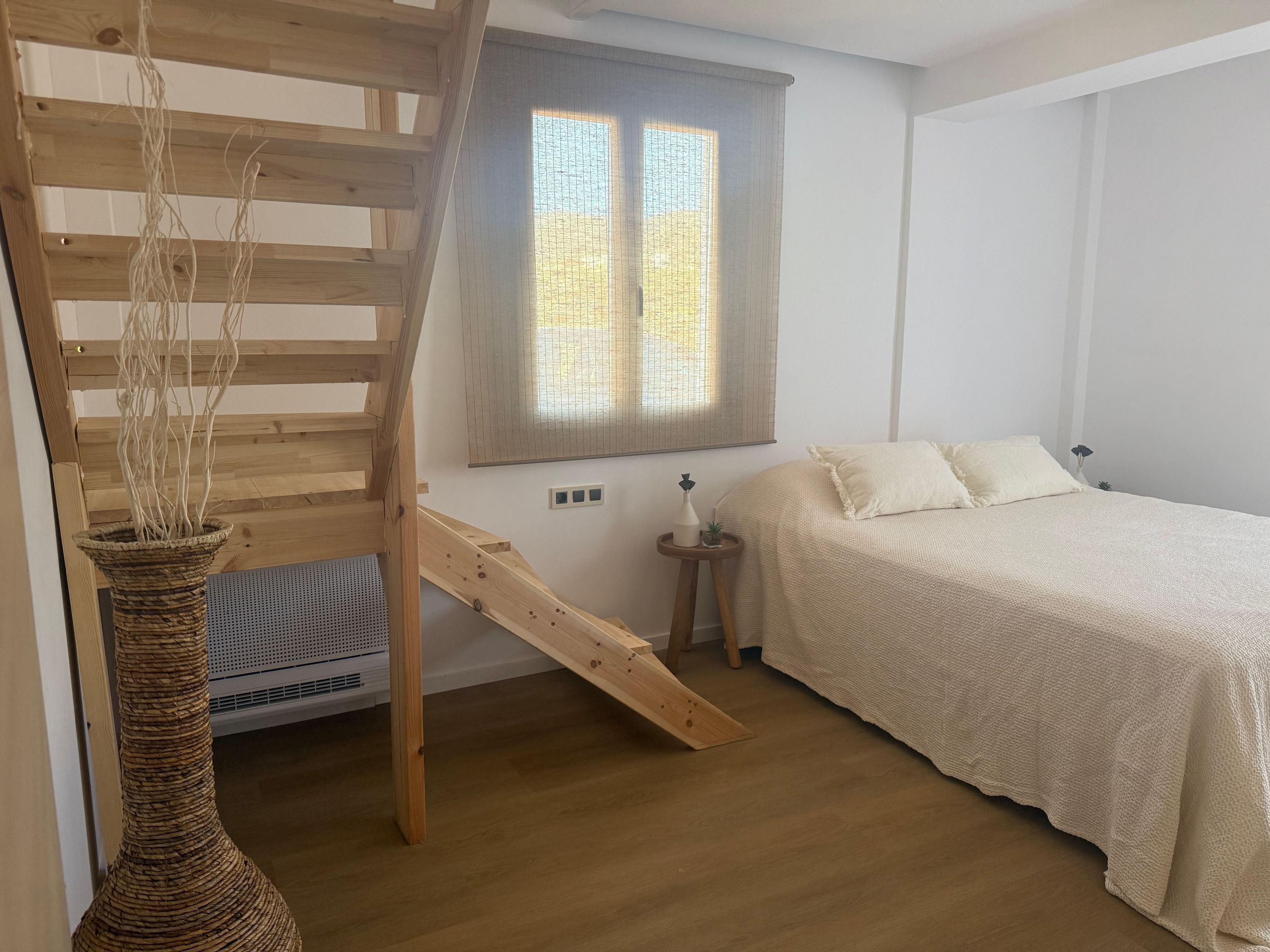 A bedroom with a wooden ladder, a bed with white bedding, a small bedside table, and a window with blinds.