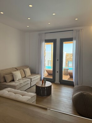 A modern living room with a sofa, a round coffee table, and a view of an outdoor area with a pool. - 2-bedroom villa Odysseus (Antiparos)
