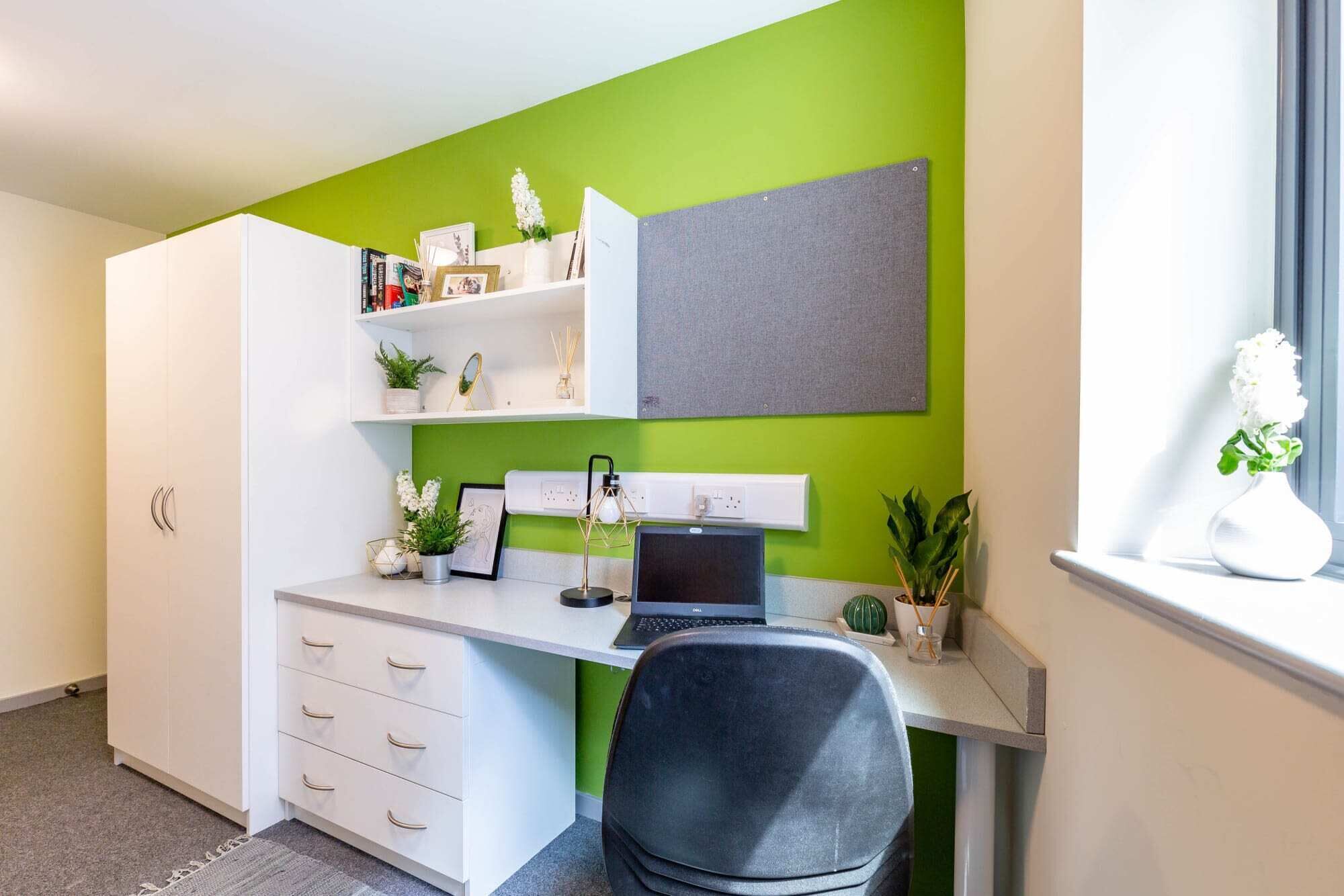 Een moderne kantoorruimte met een wit bureau, een laptop en een stoel. Er is een boekenkast met diverse spullen en een raam met een plant.