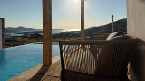Una terraza de madera con una silla de mimbre que da a una piscina y vistas al mar y colinas lejanas.
