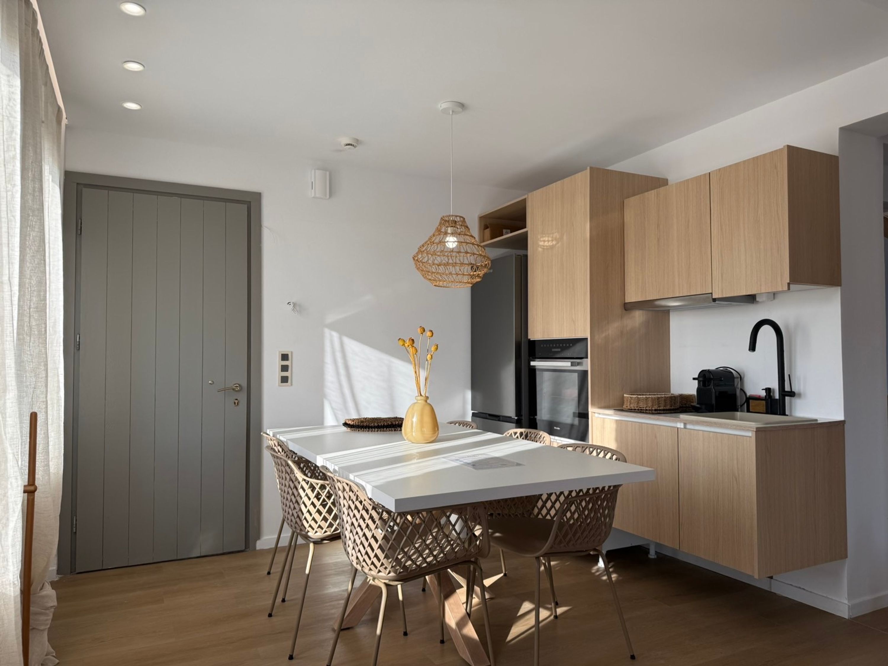 A modern kitchen with wooden cabinets, a white countertop, and a dining area with a white table and woven chairs.
