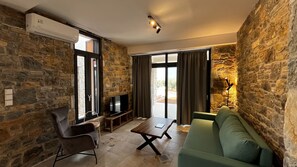 Un salon avec des murs en pierre, un canapé vert, une table basse en bois, un téléviseur posé sur un meuble et une porte-fenêtre coulissante.