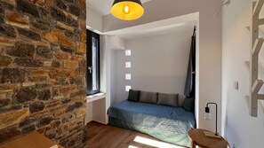 Une chambre avec un mur en pierre, un lit recouvert d’une courtepointe bleue, une fenêtre avec des stores et un luminaire suspendu.