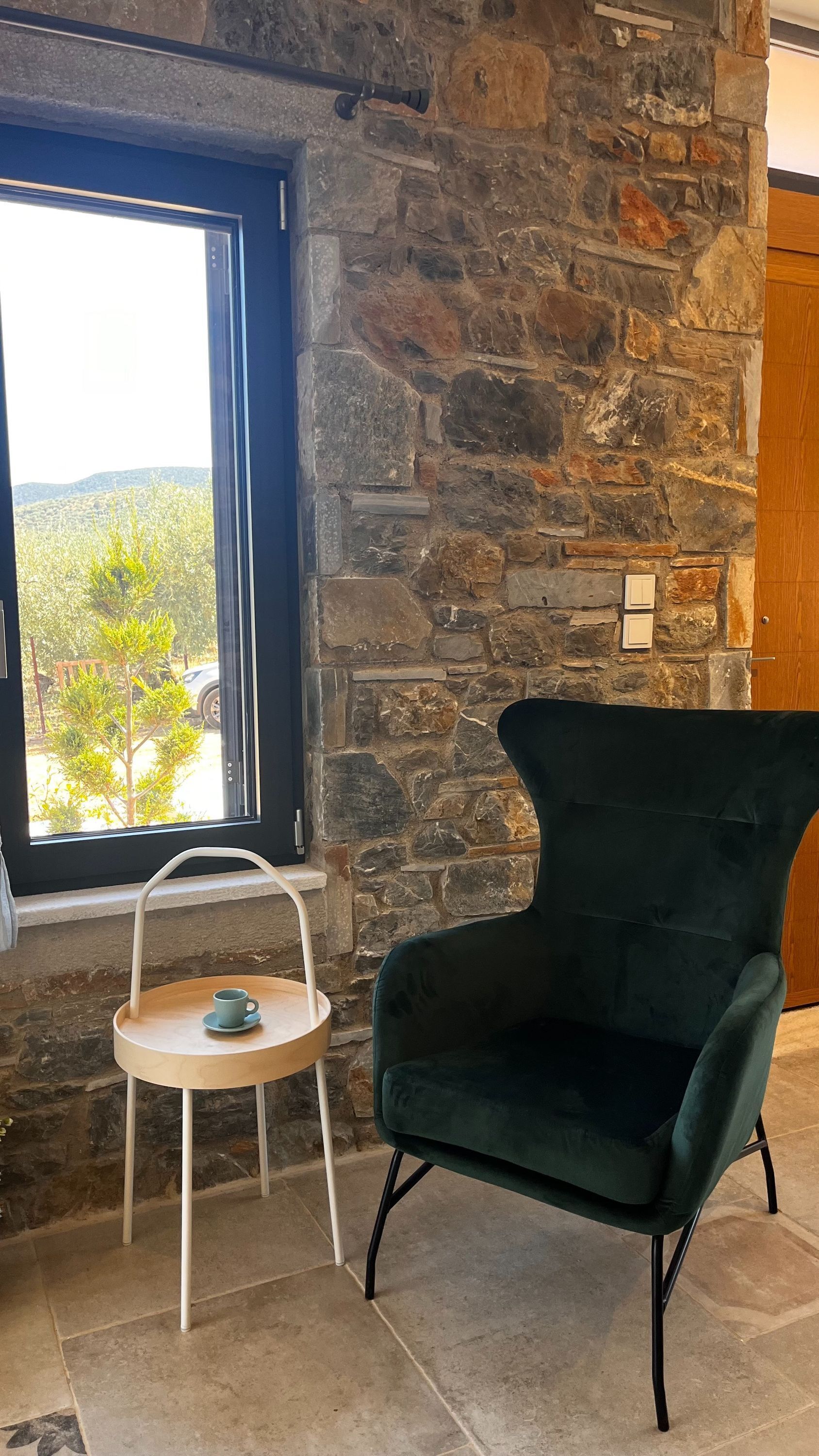 A stone wall, a window with a view of greenery and a vehicle, a dark green armchair, and a small round table with a cup.