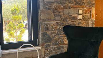 A stone wall, a window with a view of greenery and a vehicle, a dark green armchair, and a small round table with a cup.