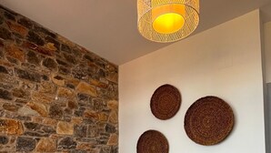 Et soveværelse med stenvæg, en seng med hvide puder, et natbord med lampe samt tre vævede vægdekorationer.