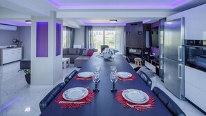 A modern dining area with a long table set for four, a kitchen with a refrigerator, and a seating area with a sofa and a TV. - DimArist Residence in Zakynthos (Agios Kirikos)