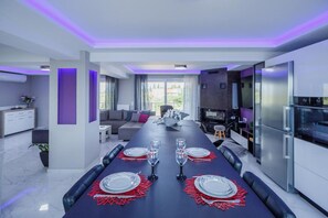 A modern dining area with a long table set for four, a kitchen with a refrigerator, and a seating area with a sofa and a TV. - DimArist Residence in Zakynthos (Agios Kirikos)