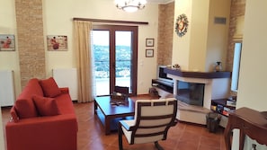 A living room with a red sofa, a wooden coffee table, a striped chair, a fireplace, and a balcony with a view. - Villa Politimi at Aegina (Egina)