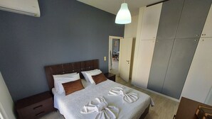 Una cama bien hecha con edredón blanco y almohadas marrones, una mesita de noche de madera, un armario con puertas correderas y una lámpara colgante.