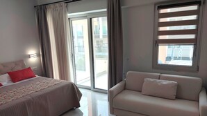 Quarto de hotel moderno com cama, sofá, janela grande com persianas e vista para a varanda externa.