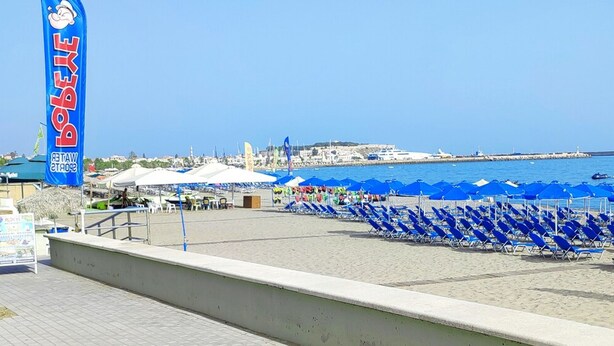 Uma praia com guarda-sóis azuis e espreguiçadeiras, um letreiro de Bobbie's, e vista para o mar e edifícios ao fundo.