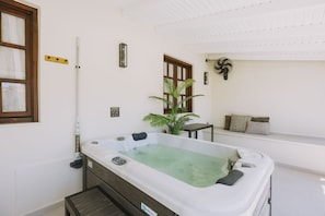 A spa room with a hot tub, a wooden bench, a potted plant, and a window with blinds. - Minares Luxury Suite (Rethymno)