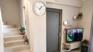 A modern living room with a staircase, a grey door, a wall-mounted clock, a flat-screen TV, and a shelf with decorative items. - Rethymno Suburb Maisonette (Rethimno)
