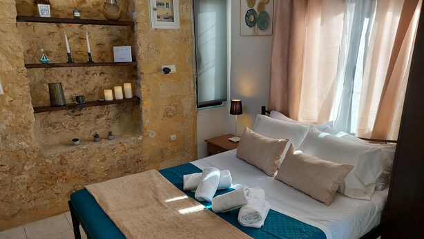 Camera d'albergo con parete in pietra, un letto con biancheria azzurra e bianca, un comodino con una lampada e uno scaffale con oggetti decorativi.