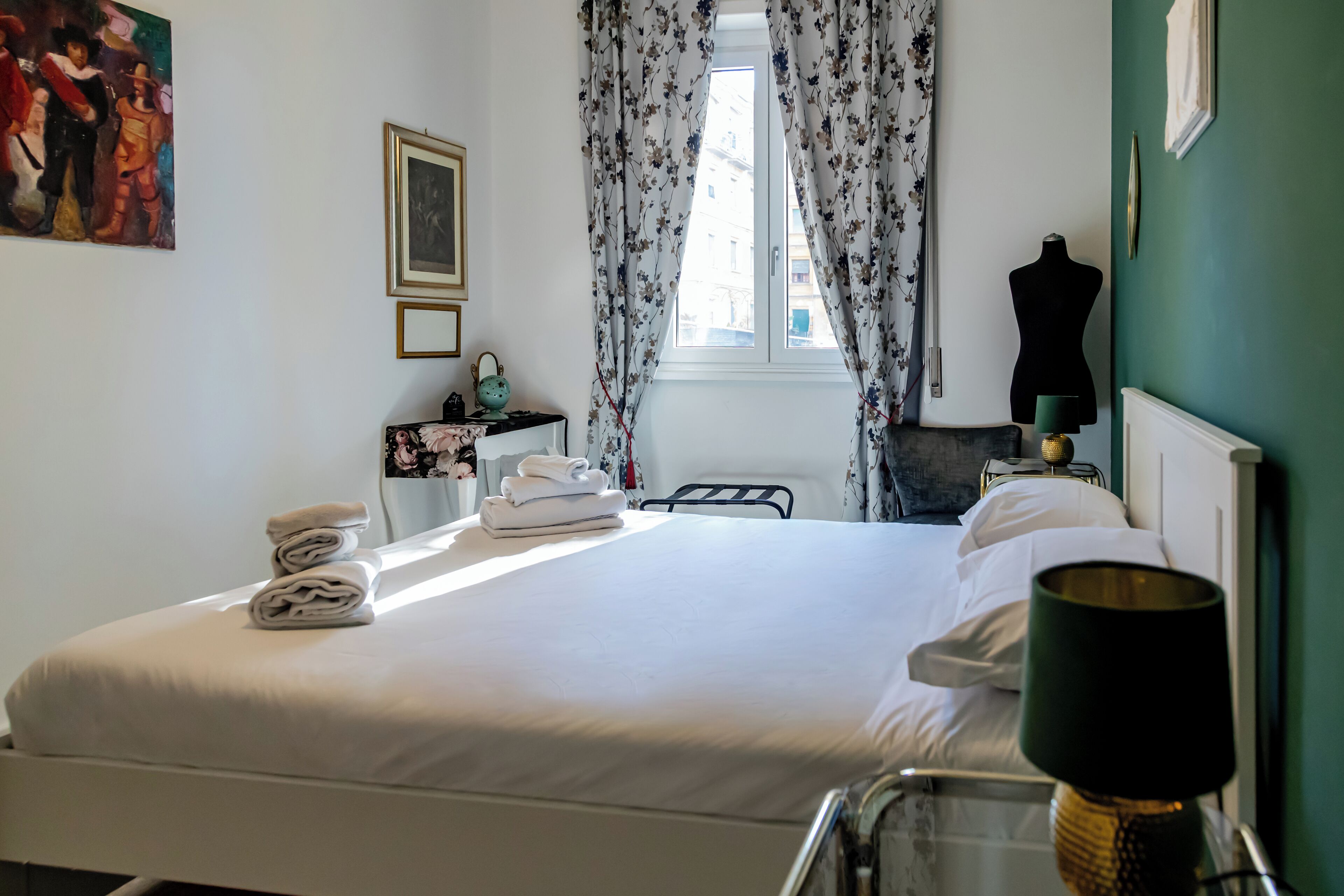 Un lit bien fait, avec des serviettes pliées, une table de chevet avec une lampe et une fenêtre ornée de rideaux à motifs floraux.