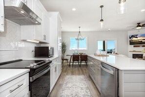 A modern kitchen with white cabinetry, stainless steel appliances, and a dining area with a wooden table and chairs. - 5BR Coastal Cottage w/ Pool, Elevator & Ocean Views built in 2022 (Emerald Isle)