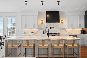 A modern kitchen with white cabinetry, a marble island, and wooden bar stools. - Oceanfront 6BR West Side Duplex w/ Pool, Elevator & Game Room (Emerald Isle)