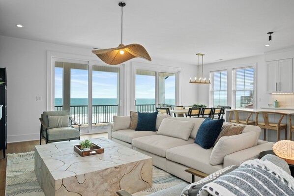 A spacious living room with a large sofa, a coffee table, and a dining area with a view of the ocean. - Oceanfront 6BR West Side Duplex w/ Pool, Elevator & Game Room (Emerald Isle)