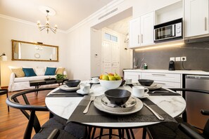 A modern dining area with a round table set for four, a white sofa with blue cushions, and a kitchen with white cabinets and a microwave. - Asty Luxury Apartments/Asty 2 (Athens)