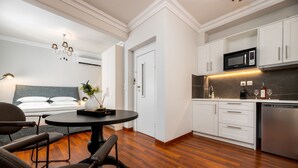Een moderne hotelkamer met een houten vloer, een ronde eettafel, een bed met een grijs hoofdeinde en een kitchenette met witte kastjes en een magnetron.