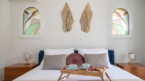 A bedroom with a large bed, two nightstands, a wooden tray with breakfast items, and two stained glass windows with wing designs. - Maio Seaview & Sunset Stay in Kremasti Rhodes (Kremasti)