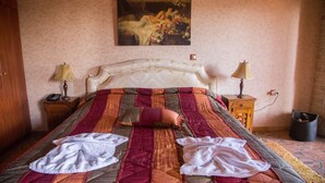 Een net opgemaakt bed met een kleurrijke quilt, twee bedlampen, een nachtkastje met lamp en een schilderij aan de muur.