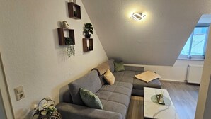 Ein modernes Wohnzimmer mit einer grauen Eckcouch, einem kleinen weißen Couchtisch und Wandregalen mit Dekorationselementen.