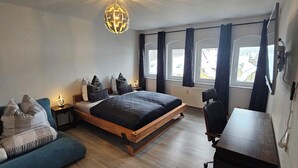 Ein modernes Schlafzimmer mit einem hölzernen Bett, einem blauen Sofa, einem Schreibtisch und einer globoartigen Pendelleuchte.
