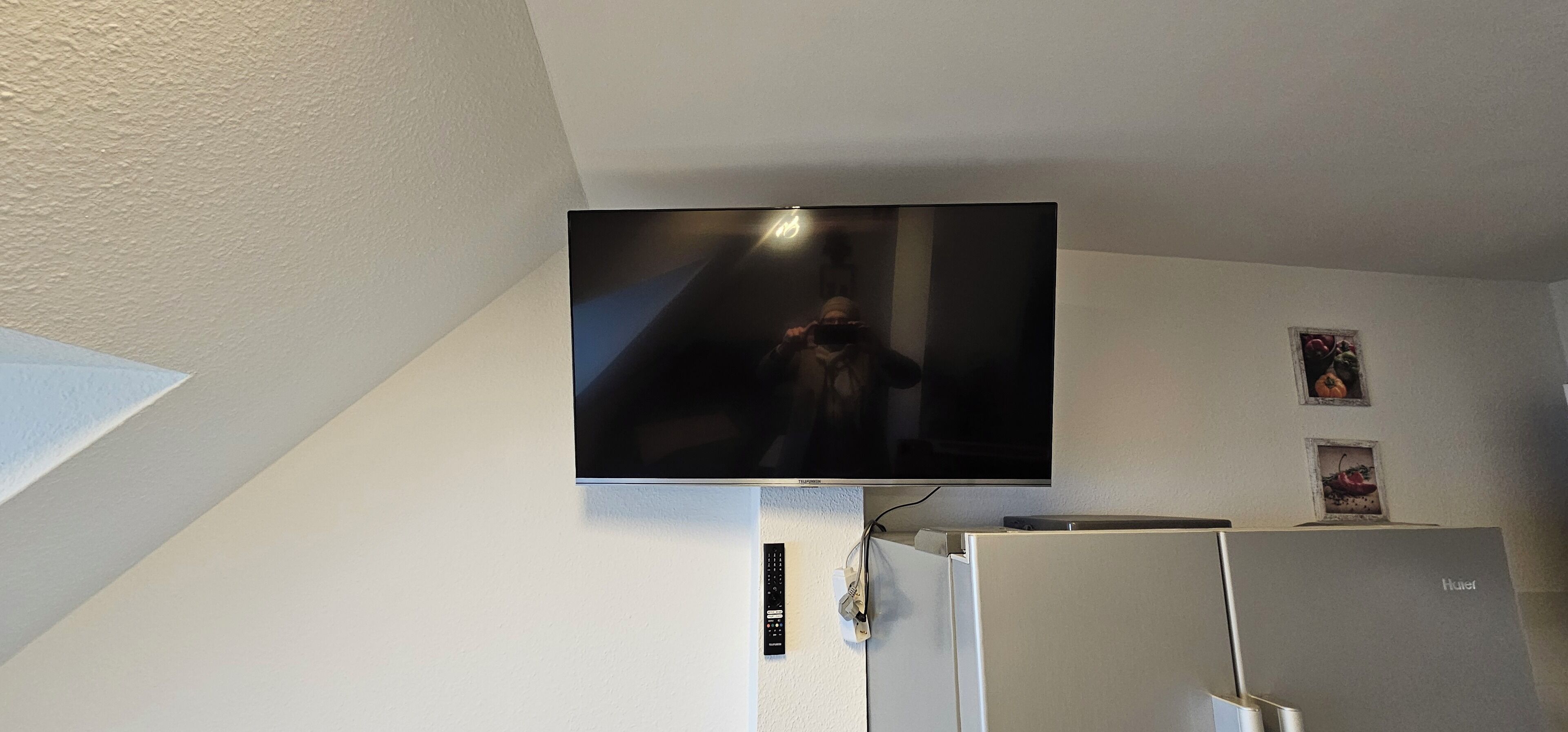 Een flatscreen televisie aan een witte muur, een koelkast en een klein, ingelijst fotootje.