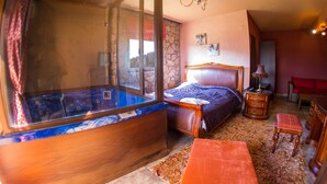 A bedroom with a large window, a bed, a wooden dresser, and a small table. - Private Jaccuzzi Suite (Kryonérion, limni plastira, karditsa)