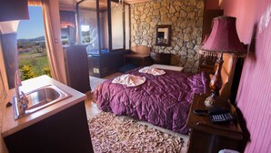 A bedroom with a stone wall, a bed with a purple bedspread, a nightstand with a lamp, and a view of the outdoors. - Private Jaccuzzi Suite (Kryonérion, limni plastira, karditsa)