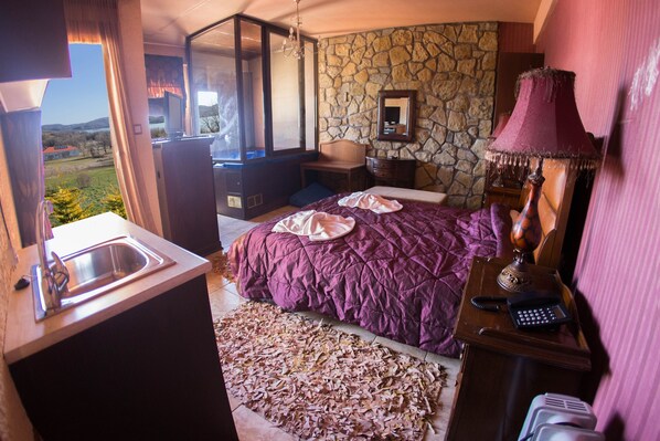 A bedroom with a stone wall, a bed with a purple bedspread, a nightstand with a lamp, and a view of the outdoors.