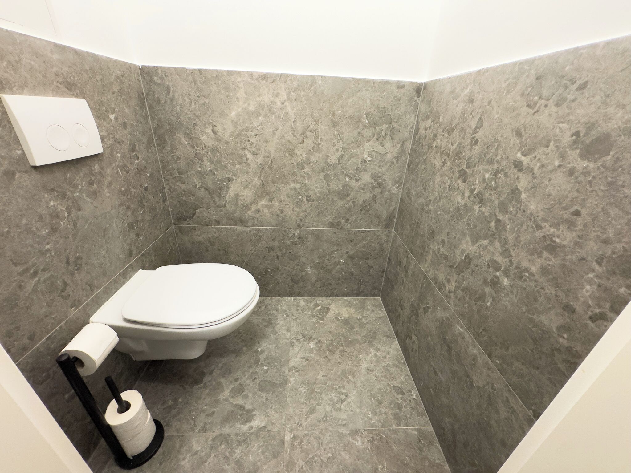 A small, modern bathroom with a toilet, a roll of toilet paper, and a black toilet brush.