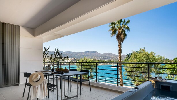Ein Balkon mit Tisch, Stühlen, einem Hut und einem Whirlpool mit Blick auf ein Gewässer und Berge.