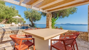 Une terrasse extérieure avec une table en pierre et des chaises en osier, donnant sur une piscine et la mer.