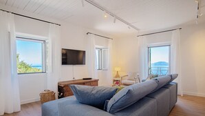 Un salon moderne avec un canapé gris, un téléviseur à écran plat fixé au mur et de grandes fenêtres offrant une vue sur la mer.