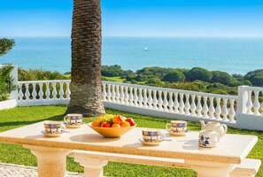 A table with a fruit bowl and tea set, overlooking a coastal view with a white fence and palm tree. - Casa Sol in Albufeira (Albufeira)